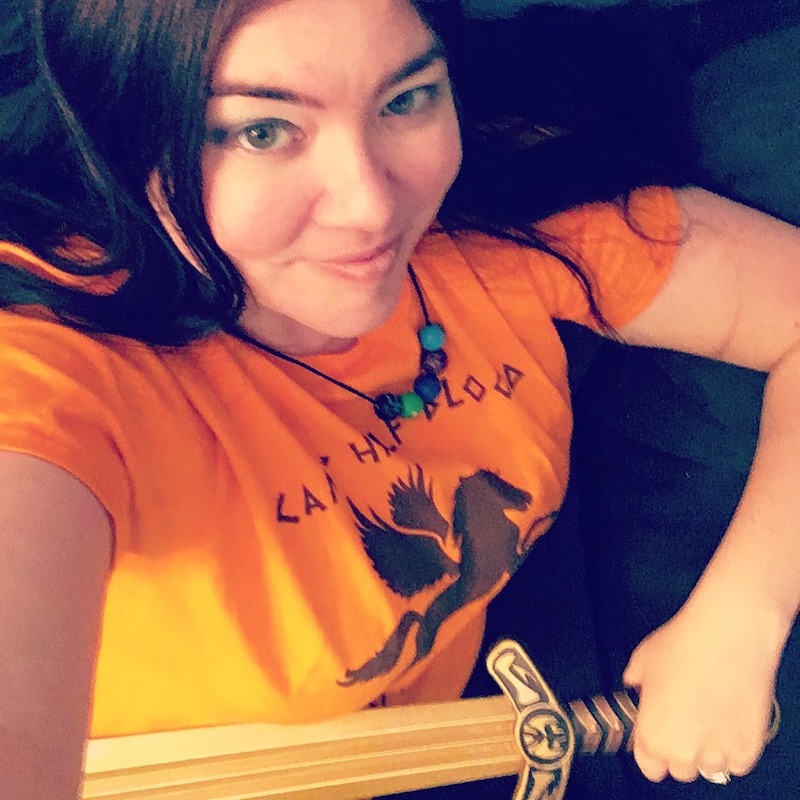 Storyteller Kat Vancil dressed in a Camp Half-Blood shirt and beaded necklace holding a replica Riptide from Rick Riordan's Heroes of Olympus storyverse